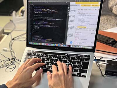 PC作業の仕事の写真。ノートパソコン上でAdobe社のDreamweaverというソフトを使ってコードを打ち込み、ウェブサイトを作成している様子。