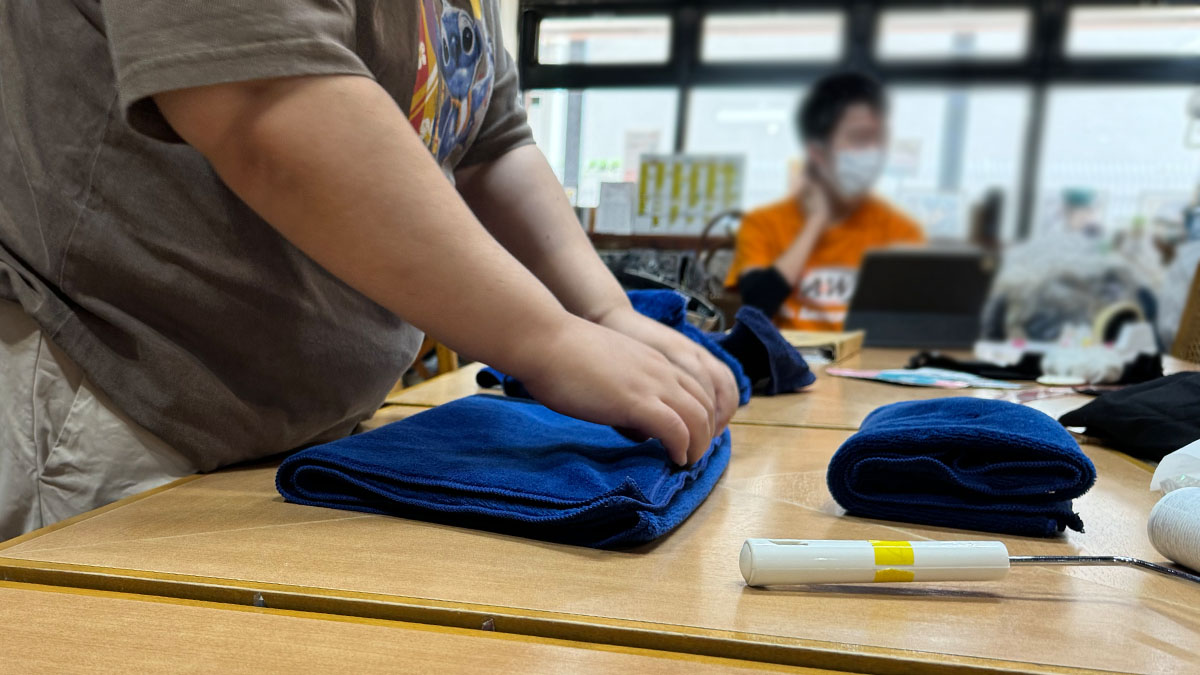 クリーニングの仕事の写真。依頼を受けて預かった青いタオルを、洗濯して干してから机に並べ、丁寧に折りたたんでいる様子。