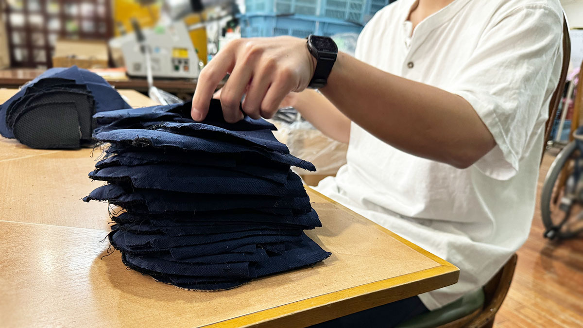 軽作業の仕事の写真。スリッパを解体している様子。二つに切って中綿を取り出したスリッパを、縫い糸を抜いて生地の形に戻し、机の上に積み上げています。